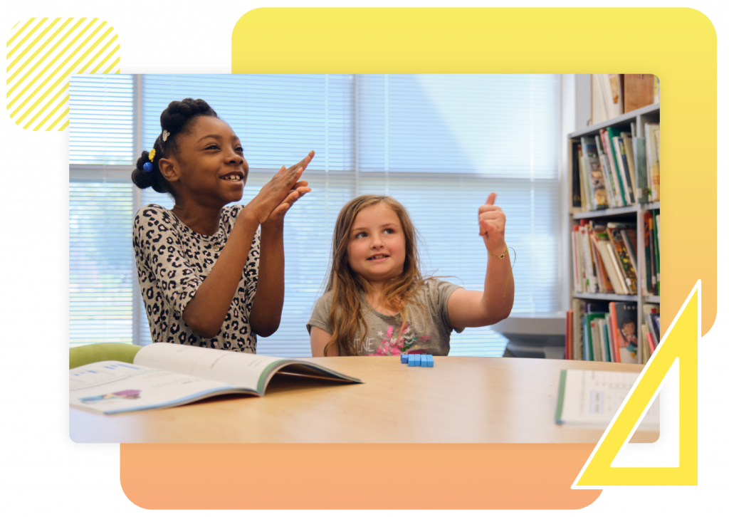 Two young girls sit at a table in a library or classroom, smiling and gesturing with their hands. Books and educational materials are on the table.