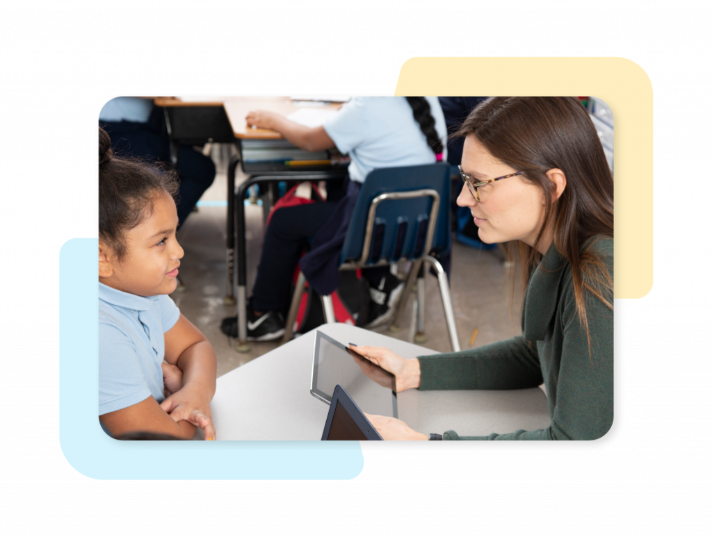 A teacher and a young student sit across from each other at a desk, talking, with a tablet on the table in a classroom setting.