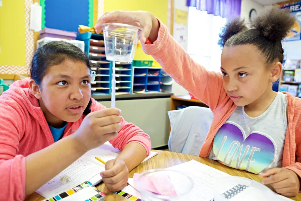 Dos estudiantes en un salón de clases realizando un experimento científico. Uno sostiene una pequeña taza transparente con líquido y una pipeta, mientras el otro observa atentamente. Los útiles escolares y los papeles están sobre el escritorio.